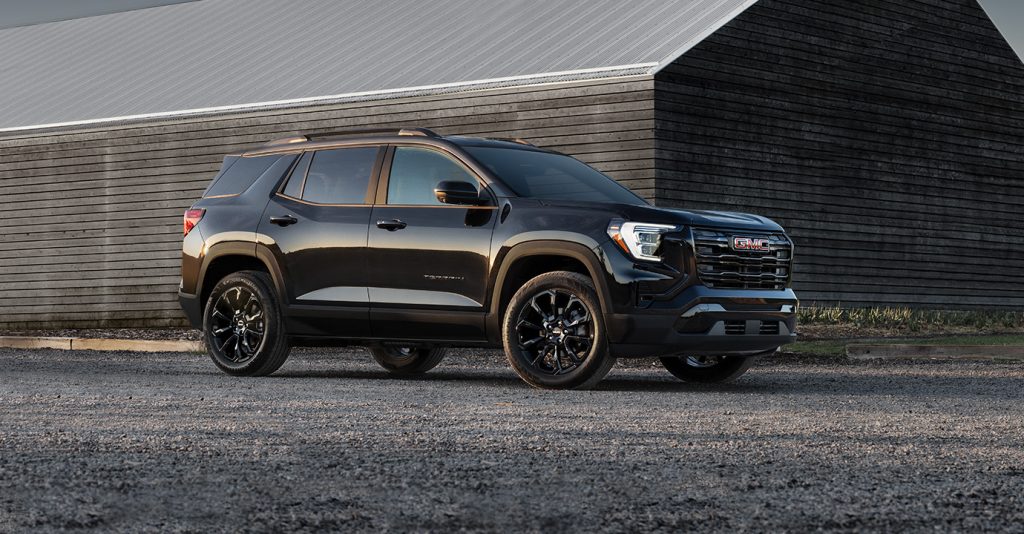 2025 GMC Terrain Elevation SUV parked on a gravel surface near a modern wooden building, featuring a sleek black exterior, bold front grille with red GMC logo, and glossy black alloy wheels in soft evening light.