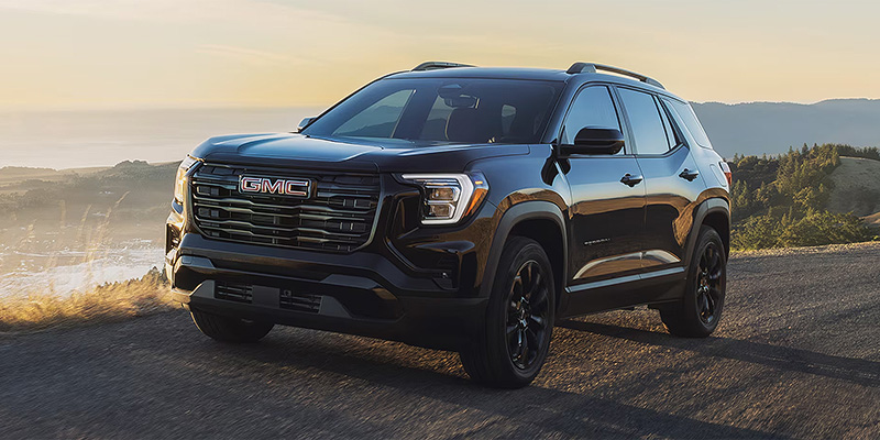 A black 2026 GMC Terrain parked on a scenic road with mountains in the background during sunset.