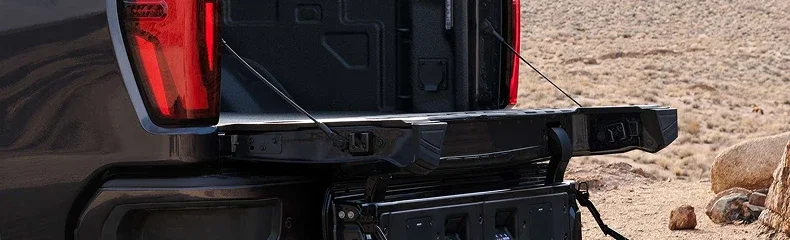 Open tailgate and rear step of a GMC Sierra HD parked on desert scrub, with the empty bed and tail lamp in view