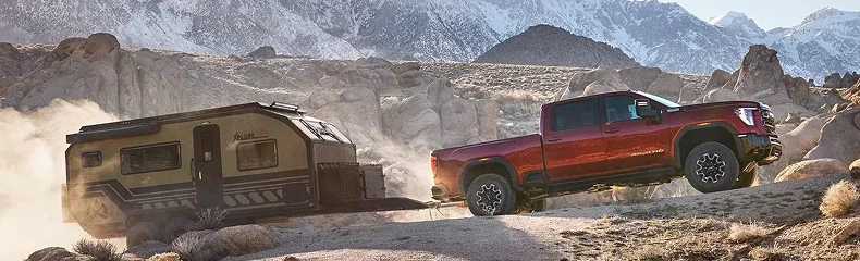 Red GMC Sierra HD towing a travel trailer along a rocky desert road with snow-capped mountains beyond
