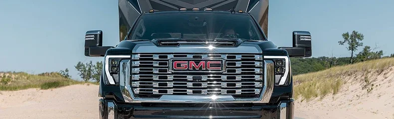 Head-on view of a GMC Sierra HD Denali chrome grille with a travel trailer hitched behind it on a sandy track