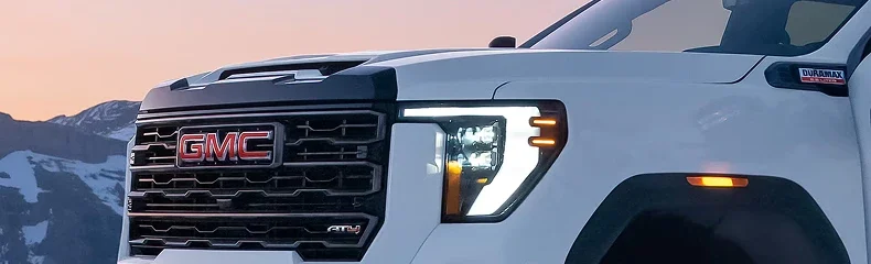 Close-up of the grille, headlamp and Duramax badge on a white GMC Sierra HD at dusk with snowy peaks behind