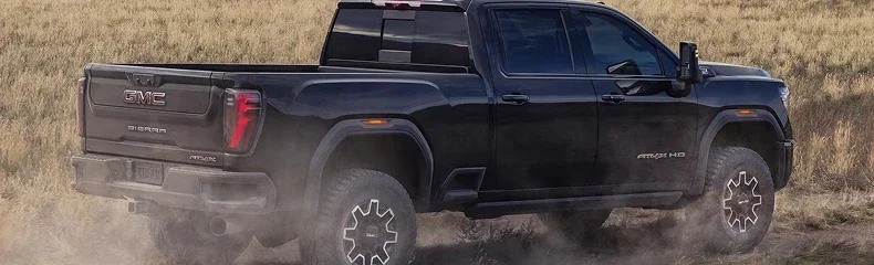 Black GMC Sierra HD AT4X seen from the rear quarter, driving away along a dusty track through flat farmland