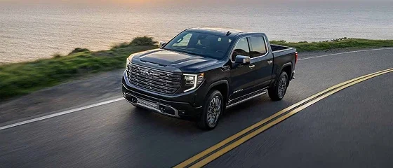 2026 GMC Sierra 1500 Denali in dark gray on a coastal highway at sunset, front three-quarter view