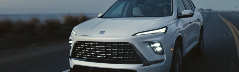 Front three-quarter view of a white Buick Enclave with its headlights on, driving along a coastal road
