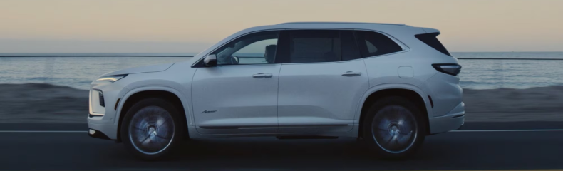Side profile of a white Buick Enclave Avenir driving along a coastal road at dusk
