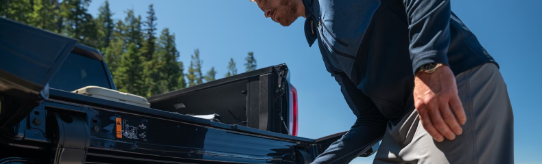 Man reaching into the open tailgate of a dark GMC pickup under a clear blue sky