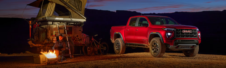 Red GMC Canyon parked at a desert campsite at dusk beside a rooftop tent and campfire